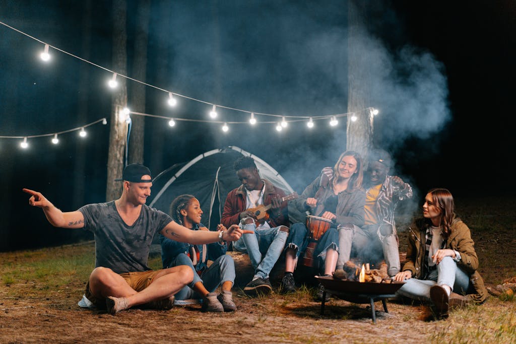 A group of friends relaxes by a campfire in the forest, playing music and talking.