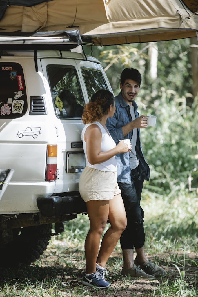 Side view of positive romantic young campers in casual clothes leaning on trunk of SUV car and drinking coffee during trip in forest on sunny summer day
